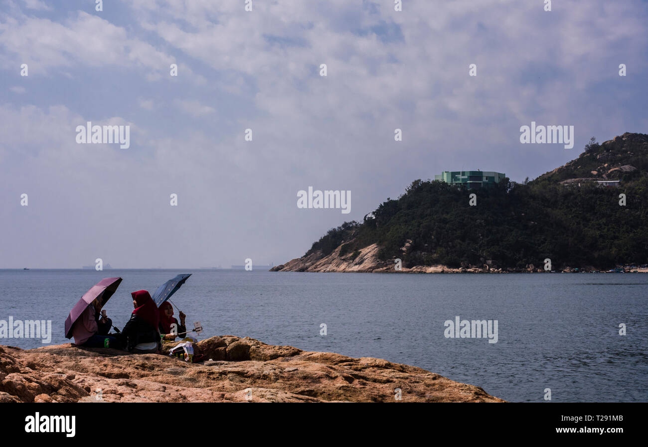 Drei Leute sitzen auf den Felsen, mit Sonnenschirmen, sich von der Sonne abzuschirmen, im Küstenort Stanley, Hong Kong Stockfoto