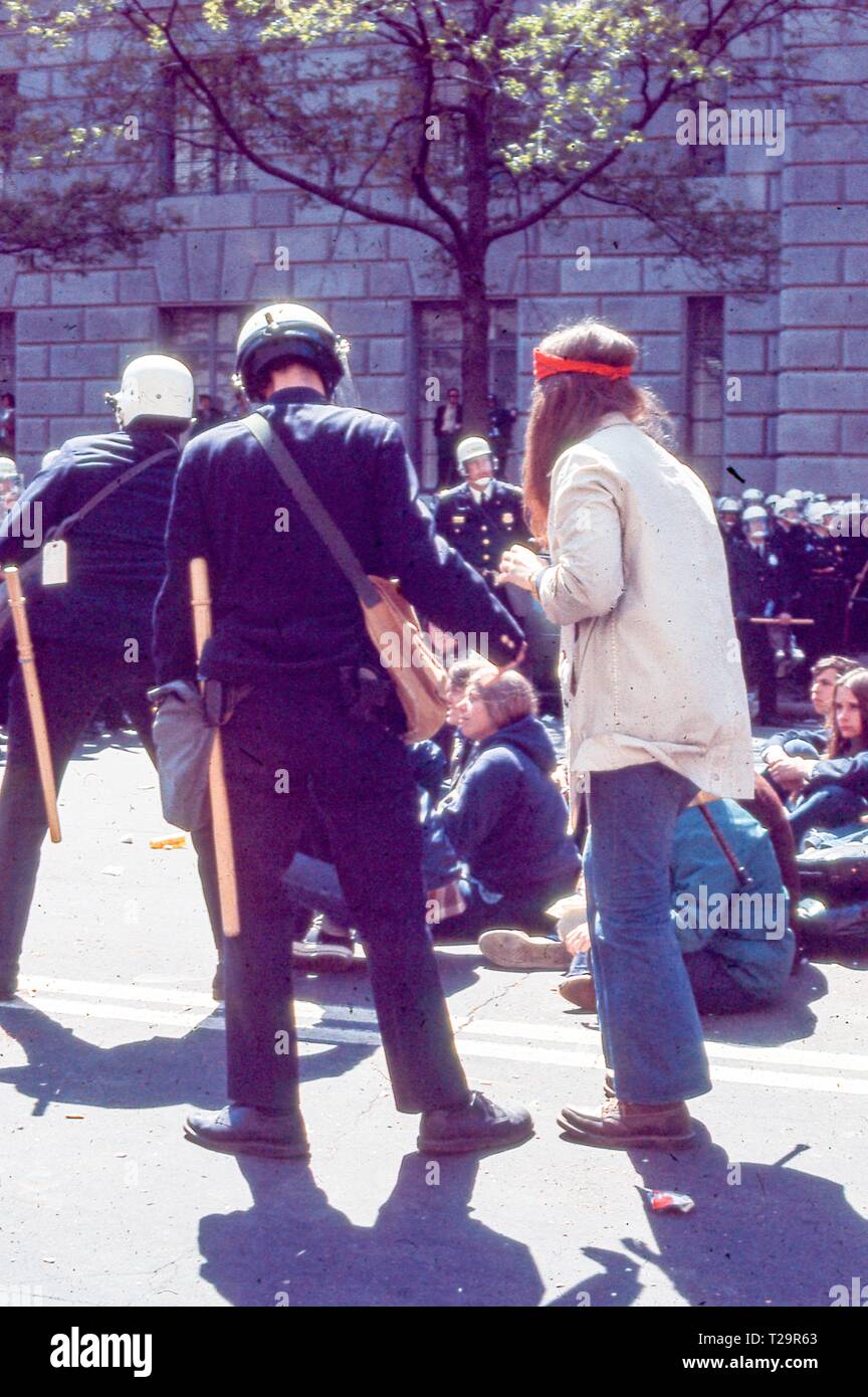 Während der 1971 Tag der Proteste gegen den Vietnamkrieg, eine männliche Demonstrant mit langen dunklen Haaren steht in voller Länge Profil in der Nähe der Zahlen von zwei Polizisten (beide von der Kamera), die blauen Uniformen in der Nähe des Internal Revenue Service Gebäude, in Washington, DC, Mai 1971 tragen. () Stockfoto