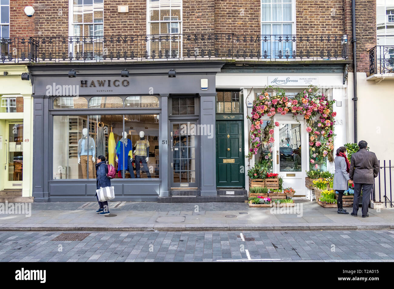 Eine Frau geht an Hawico vorbei, einem luxuriösen Kaschmir- und Strickladen, während ein Paar sich die Ausstellungen außerhalb der Blumenhändler von Moyses Stevens in Belgravia, London, Großbritannien, ansehen kann Stockfoto