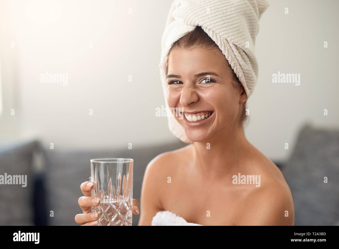 Gerne attraktive junge Frau mit einem netten freundlichen Grinsen tragen ein sauberes weißes Tuch um Ihr Haar halten ein Glas frisches Wasser am cam Lachen Stockfoto