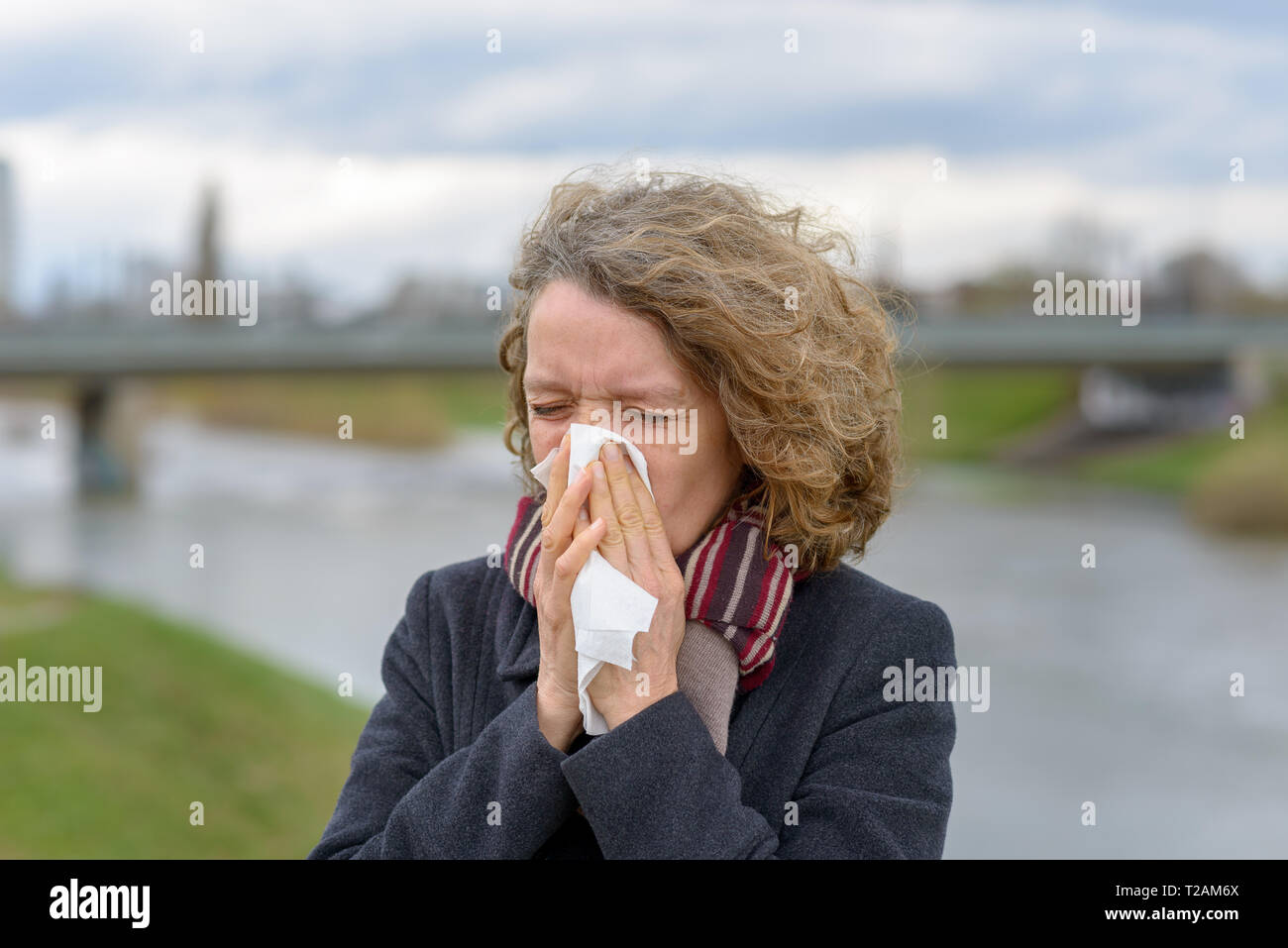 Frau trägt einen warmen Winter Mantel und Schal ihre Nase weht auf ein Gewebe im Freien in einer kalten, bewölkten Tag in ein Konzept der kranken Gesundheit, Erkältungen und Grippe oder r Stockfoto