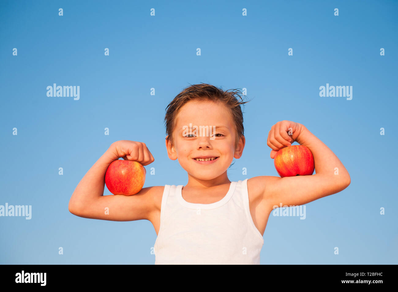 Glücklichen kleinen kaukasischen Kind in weißes Tank Top mit Muskel Bizeps mit Äpfeln auf blauen Himmel Hintergrund Stockfoto