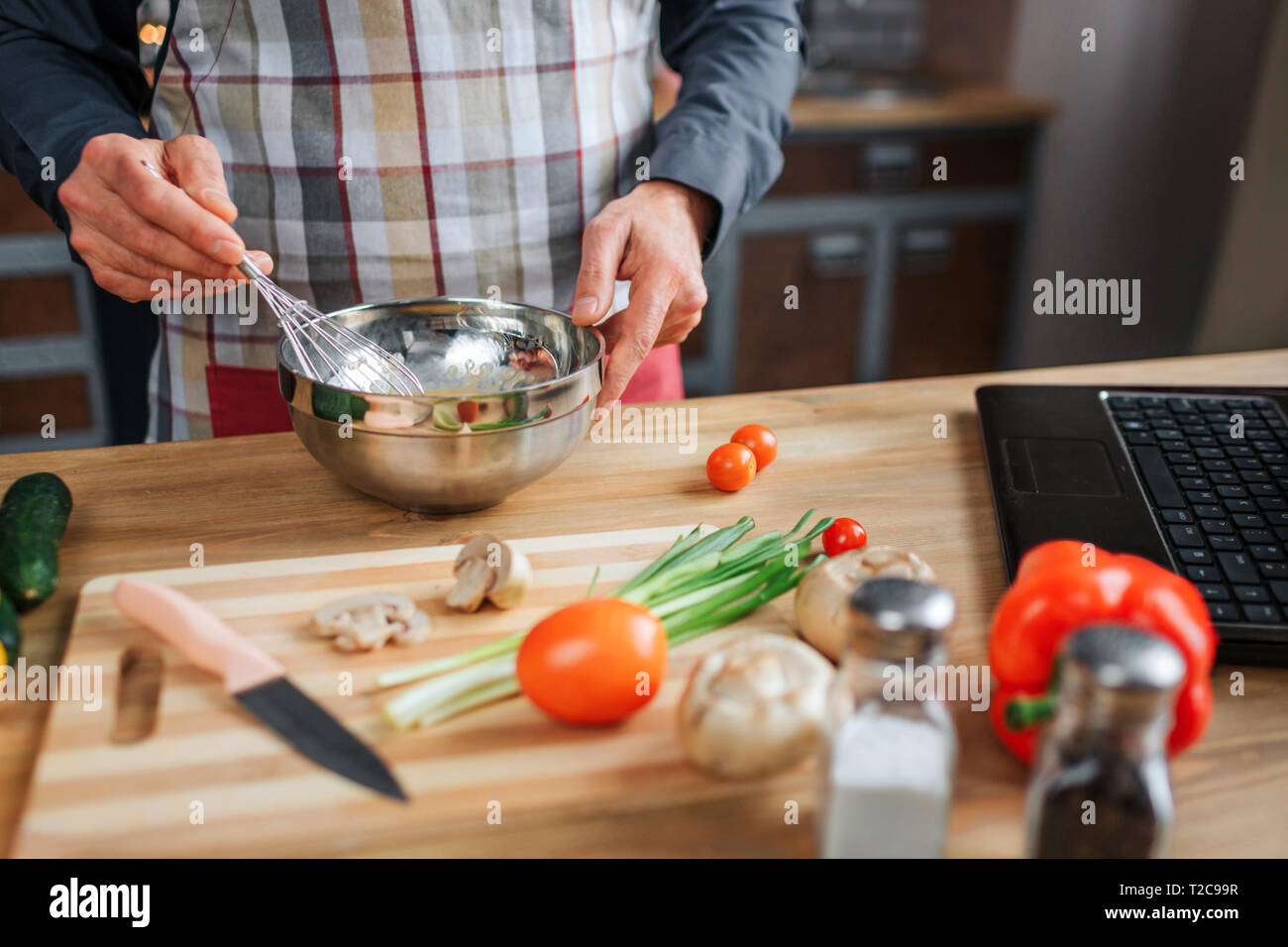 In der Nähe der Hände des Menschen mischen Eier in der Schale. Er arbeit am Tisch in der Küche. Guy tragen Schürze. Buntes Gemüse und Laptop Tastatur auf dem Schreibtisch Stockfoto