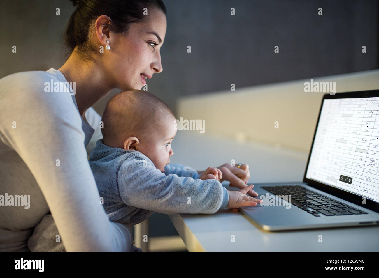 Eine Seitenansicht eines jungen Studenten Mutter oder Geschäftsfrau, sitzen am Schreibtisch mit einem Baby im Zimmer in einer Bibliothek oder einem Büro, mit Laptop. Stockfoto