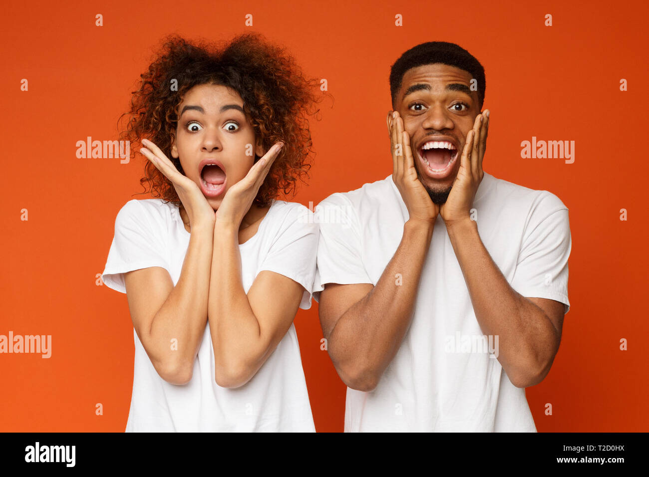Schwarzer Mann und Frau Eröffnung Mund erstaunt und berührt, Wangen auf Orange Studio Hintergrund Stockfoto