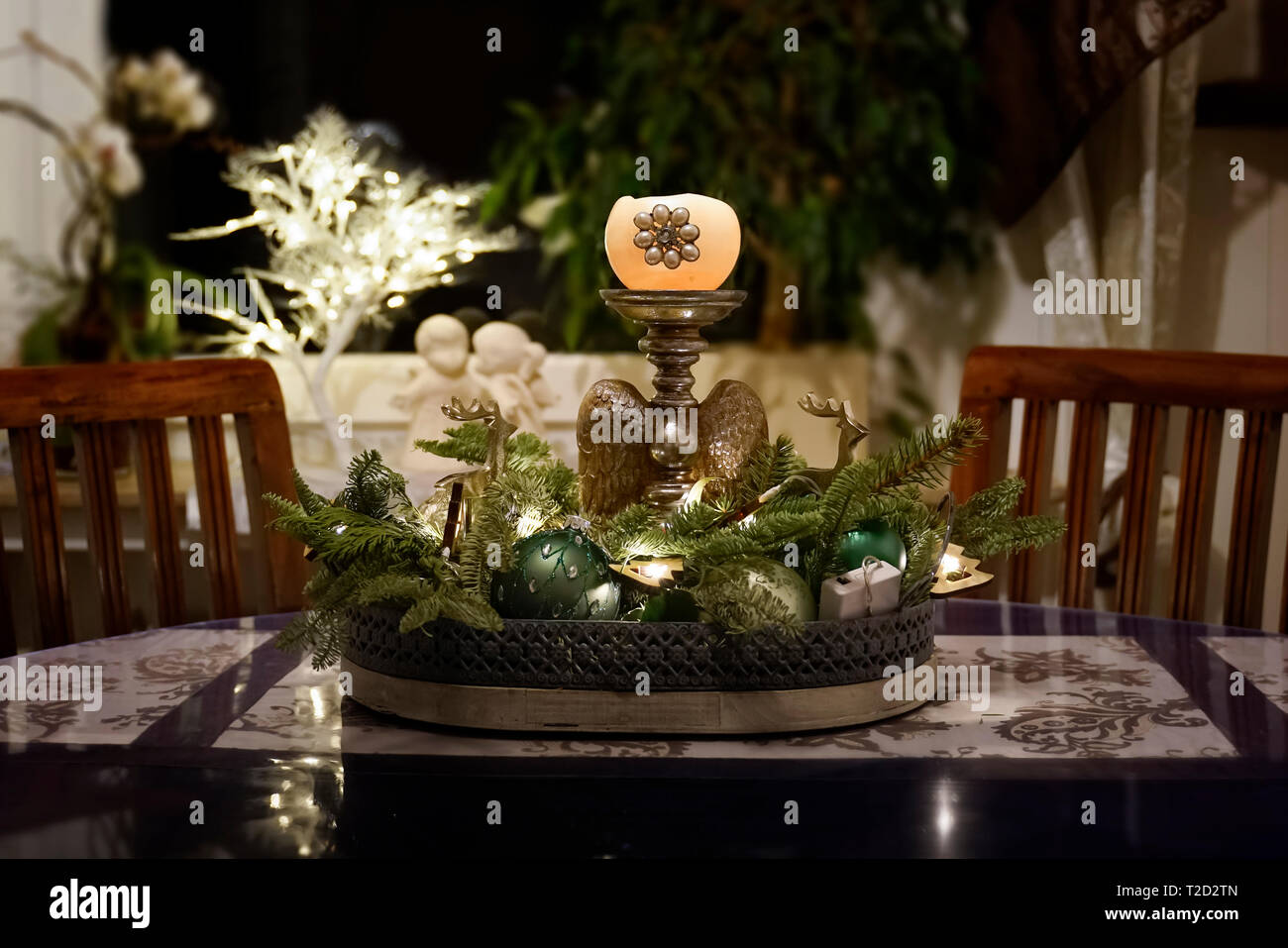 Eine Anordnung mit Kerze und grünen Tannenzweigen schmückt ein Zimmer zur Weihnachtszeit. Dunkle Holzmöbel und ein Fenster mit Gips Engel und Illumina Stockfoto