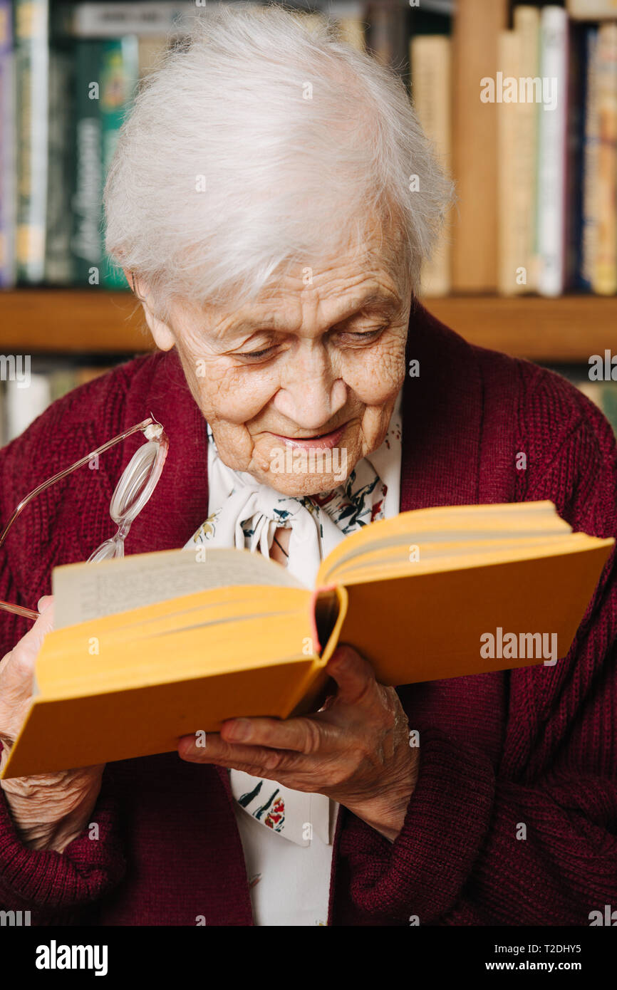 Portrait von fröhlichen älteren grau behaarte Frau. Sie liest, stehend mit Buch in der einen Hand und Gläser in einem anderen, eng gebogen siehe Buchstaben bett Stockfoto