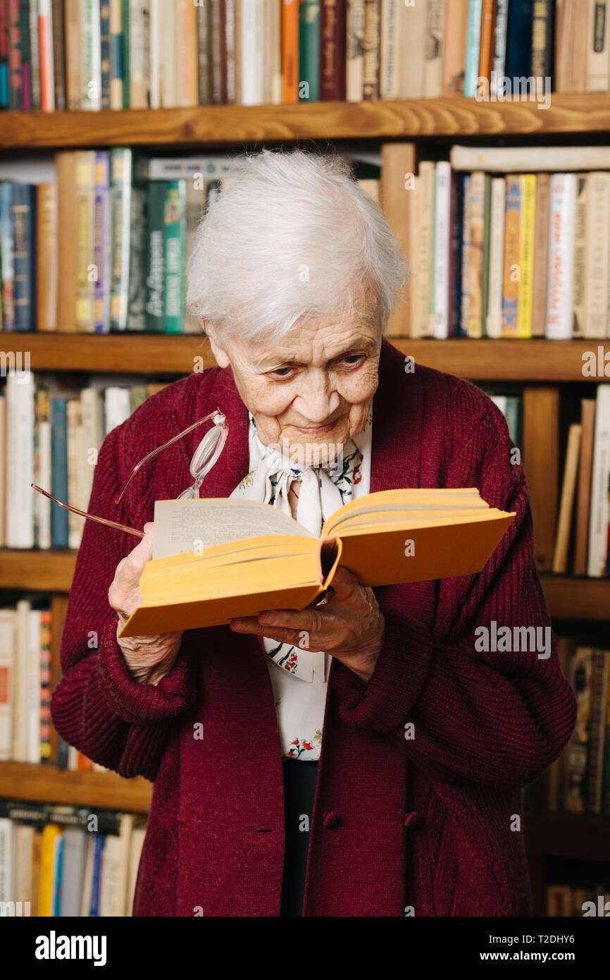 Portrait von fröhlichen älteren grau behaarte Frau. Sie liest, stehend mit Buch in der einen Hand und Gläser in einem anderen, eng gebogen siehe Buchstaben bett Stockfoto