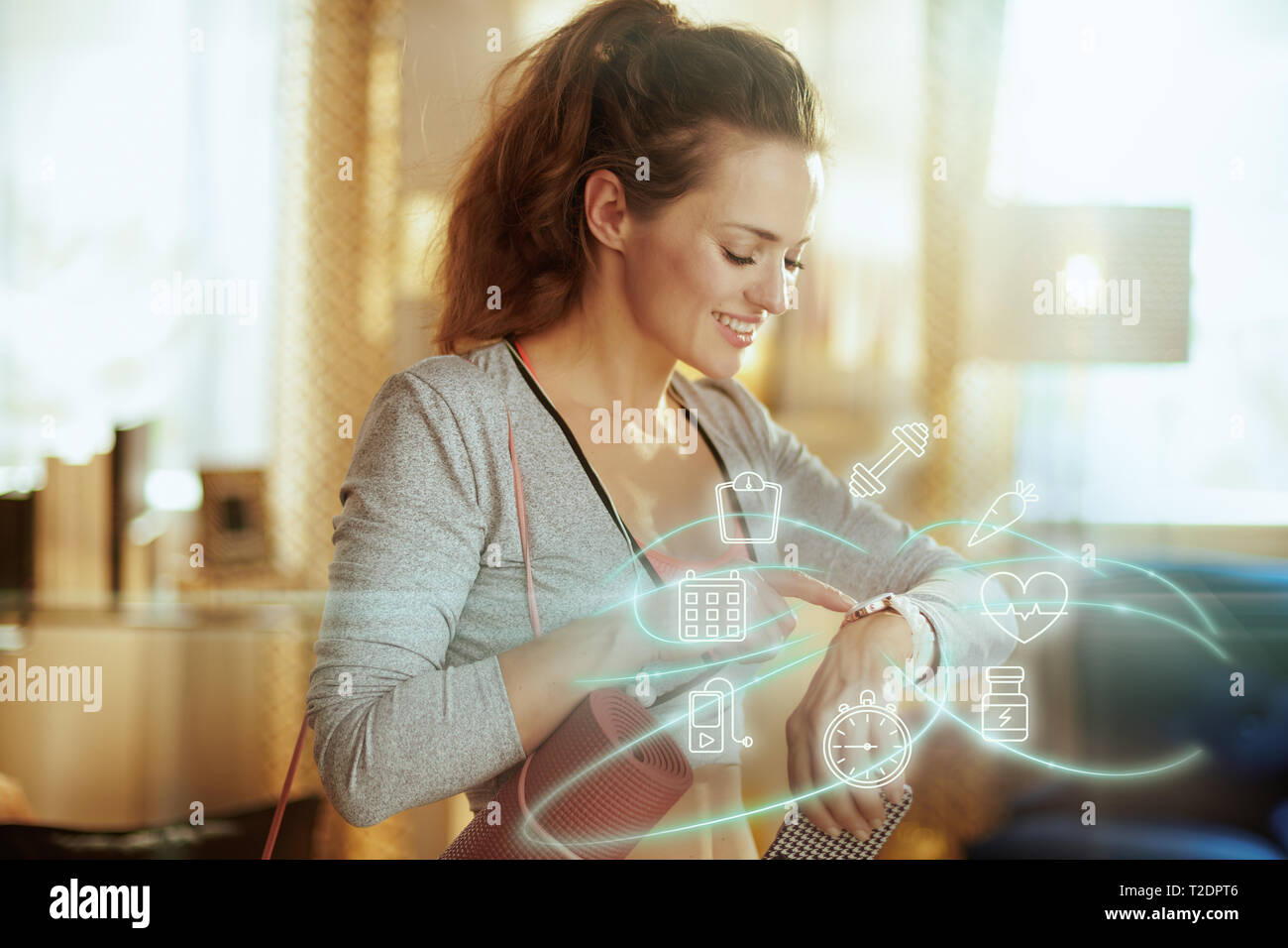 Lächelnd passende Frau im Sport Kleidung mit Fitness Matte mit smart Watch in das moderne Haus zu verfolgen, fährt oder reitet in Fitness app. Stockfoto