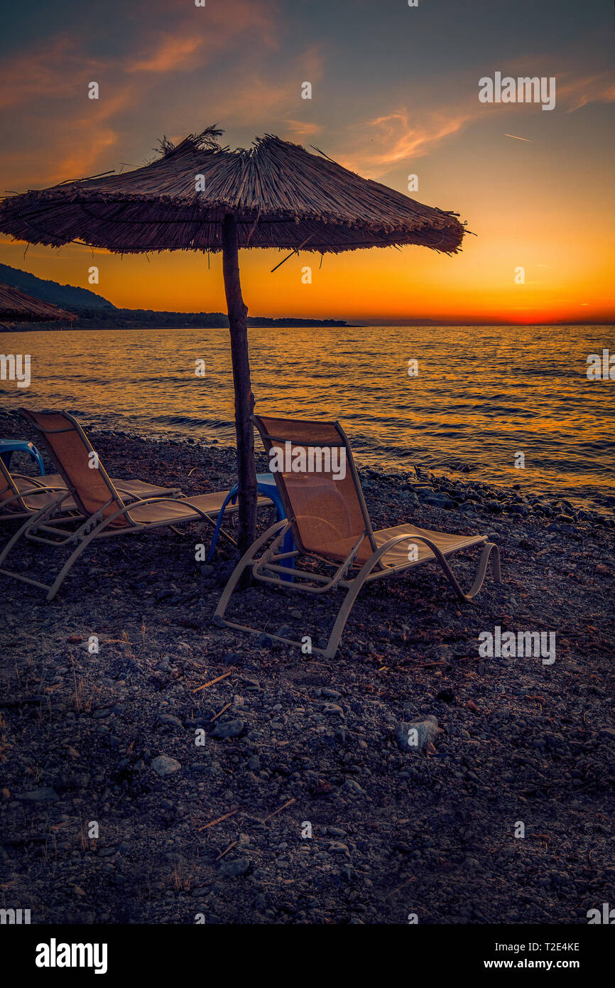 Sonnenliegen auf einer felsigen Strand bei Sonnenuntergang mit einem Stroh Regenschirm perfekt für Touristen auf der Insel Samothraki in Griechenland mit einer lebendigen Sky Stockfoto