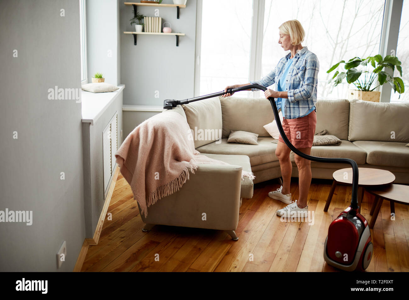Schwerwiegende attraktive reife Frau in homewear mit Staubsauger reinigen Polster auf dem Sofa im Wohnzimmer. Stockfoto