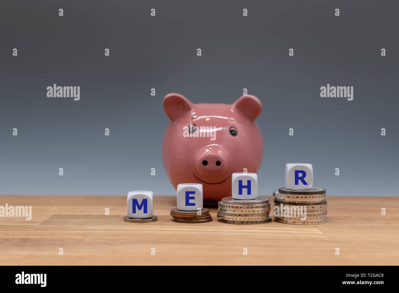 Symbol für mehr Geld. Würfel auf Stapel von Münzen aus dem deutschen Wort 'mehr' (Erz" in englischer Sprache). Ein sparschwein ist im Hintergrund. Stockfoto