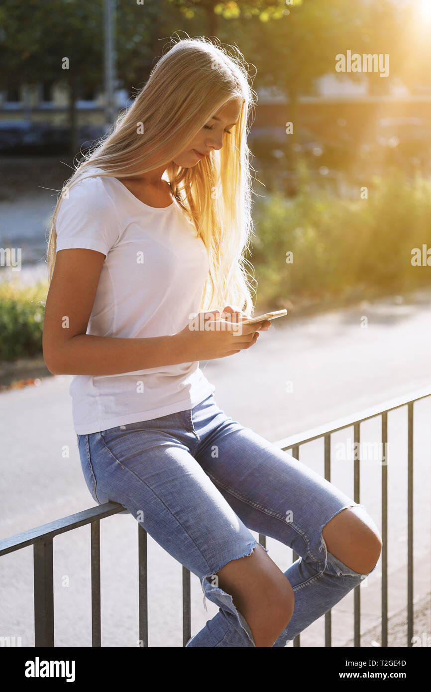 Urban teenage Mädchen mit ihrem Smartphone oder Handy beim Sitzen auf Geländer von city street - ehrliche reale Menschen Technologie Konzept - Hintergrundbeleuchtung wi Stockfoto