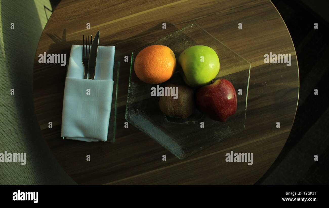 Ein Glas Schale mit Apfel, grüner Apfel, Orange und Kiwi, mit Messer und Gabel in Serviette auf einem Holz- Runder Tisch Stockfoto