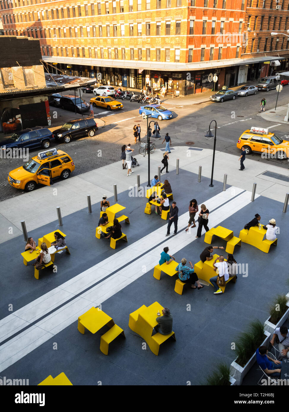 Bei dieser Hochwinkelaufnahme werden Menschen in einem öffentlichen Außenbereich um gelbe Tische sitzen und stehen gesehen. Die Szene spielt in einer Stadt mit Gebäuden und Straßen im Hintergrund. Die Leute genießen wahrscheinlich den öffentlichen Raum, vielleicht essen, plaudern oder einfach nur entspannen. Die gelben Tabellen verleihen der Szene einen Farbakzent und machen sie optisch ansprechend. Stockfoto