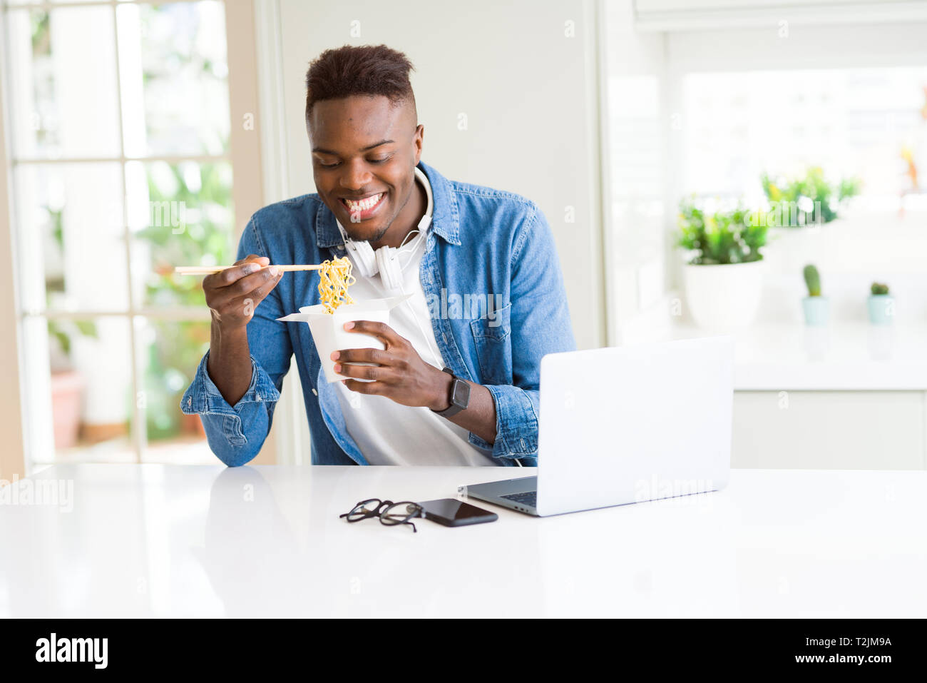 Hübscher junger African Business Mann essen Lieferung asiatische Lebensmittel und Arbeiten mit Computer, genießen Nudeln lächelnd Stockfoto