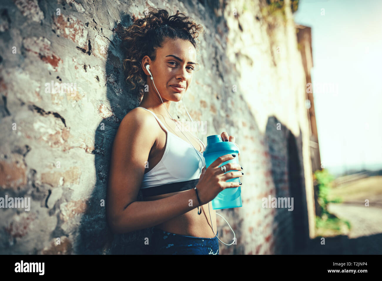 Eine junge muskulöse Frau, mit Kopfhörer, steht gegen die Wand und Trinkwasser nach der Ausbildung. Stockfoto