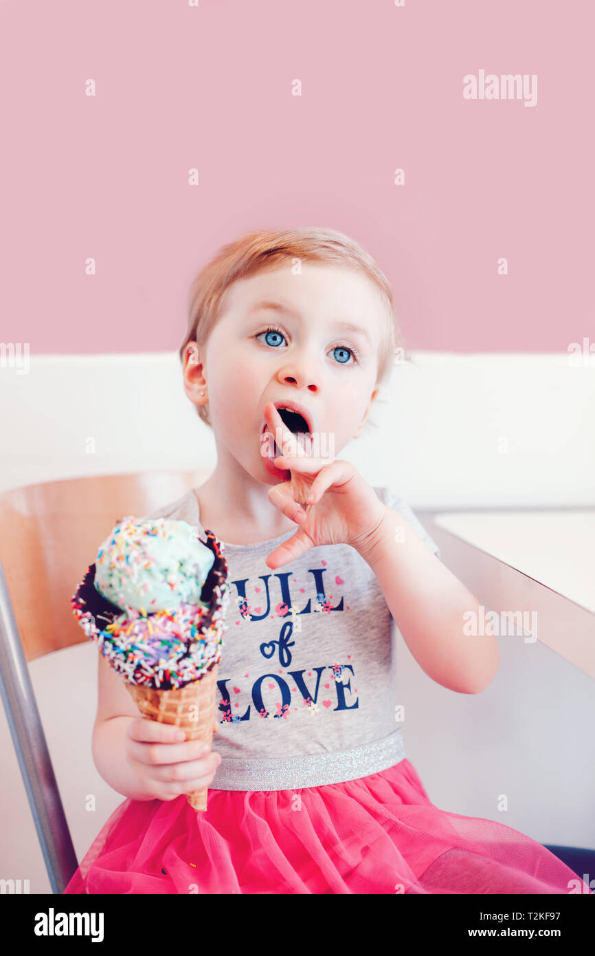 Cute adorable lustig Kaukasischen blond babyl Mädchen mit blauen Augen Eis essen in große Waffel Kegel mit bunten Streuseln. Kid lecken Finger Stockfoto
