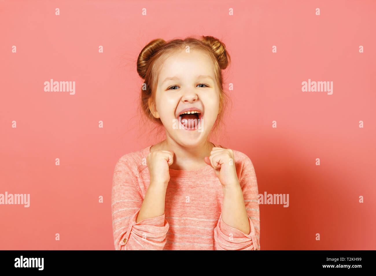 Closeup Portrait von einem netten attraktiven fröhliche kleine Mädchen mit Brötchen in Ihr Haar über rosa Hintergrund. Das Kind öffnete seinen Mund und hält seine Fäuste. Stockfoto