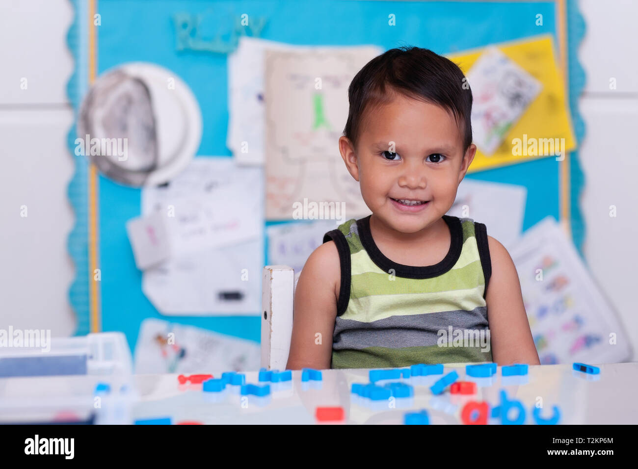 Ein niedliches Hispanic junge sitzt in der Kindertagesstätte mit Alphabet Buchstaben auf dem Tisch und einer bunten Bulletin Board im Hintergrund. Stockfoto