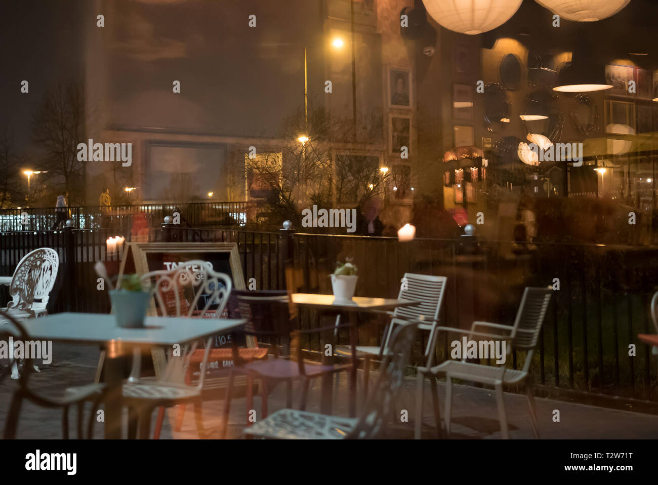 Blick von innen auf die trendige Bar/Bistro/Cafe, abends von außen auf der Straße. Stühle und Tische im Freien, die durch die Spiegelung des Fensters sichtbar sind. Stockfoto