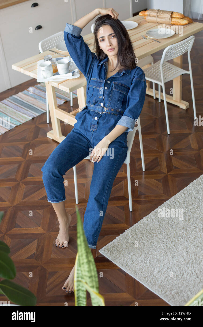 Schöne Frau in Jeans Overalls sitzt auf einem Stuhl in der Küche  Stockfotografie - Alamy
