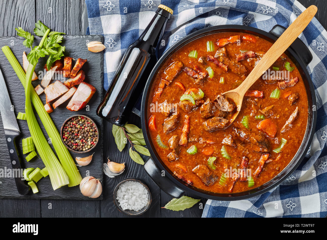 Traditionelle irische Rindfleisch und Bier Eintopf mit Karotten, Sellerie, Karotten und Gewürze in einem holländischen Ofen mit Zutaten und Flasche mit Bier auf einem schwarzen w Stockfoto