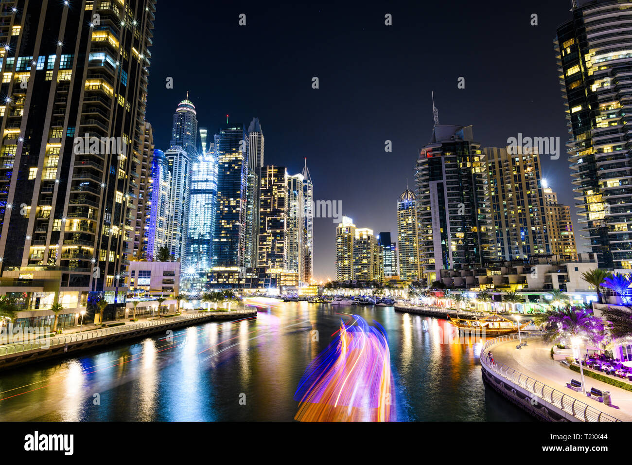 Atemberaubende Aussicht auf den Jachthafen von Dubai in der Dämmerung mit beleuchteten Wolkenkratzer im Hintergrund und einer leichten Wanderwegen durch eine Yacht segeln im Vordergrund links. Stockfoto