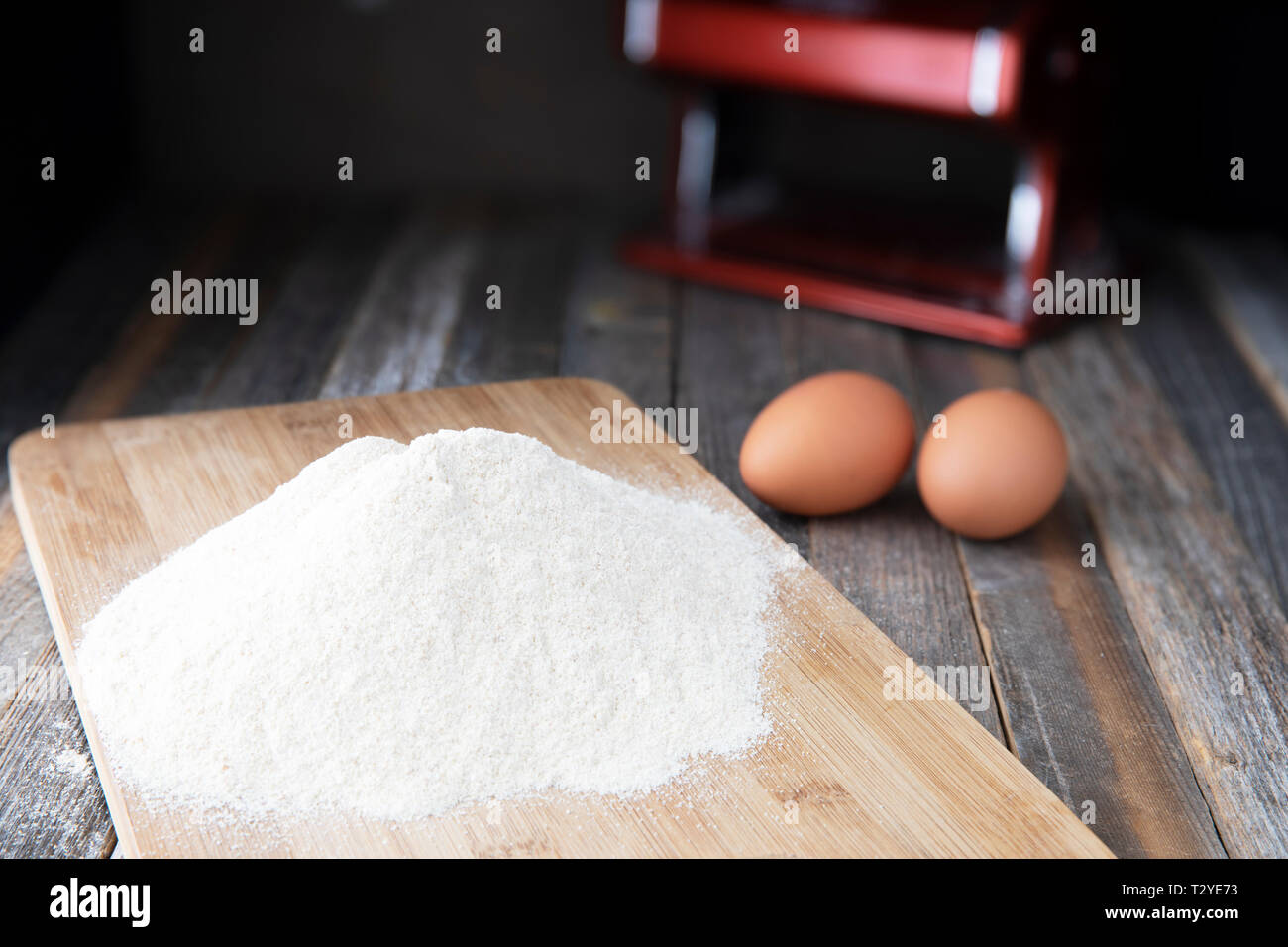 Mehl auf ein Schneidbrett mit zwei Eiern und pasta Maschine im Hintergrund. Stockfoto