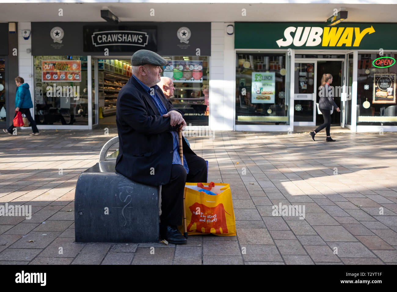 Ältere Mann in Tuch cap. mit Stock und Shopping Bag, sitzend auf einer konkreten Bank auf dem Moor Fußgängerzone in Sheffield Stockfoto