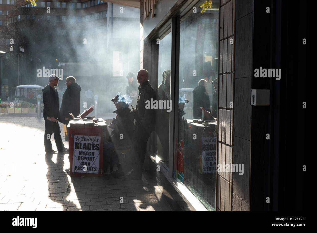 Mann vaping durch einen Kiosk an der Straßenecke in Sheffield, mit Rauch- und Passanten durch die Sonne Silhouette Stockfoto