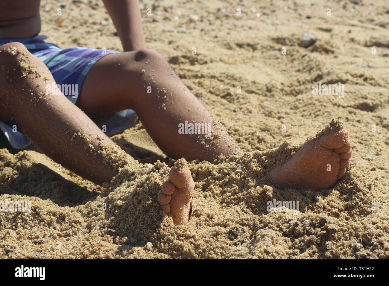 Junge in Shorts mit Beinen teilweise in Sand am Strand bedeckt Stockfoto