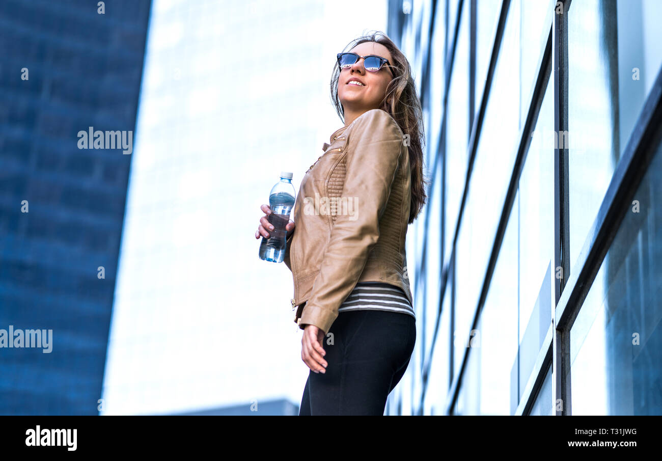 Selbstbewusste Frau in der Stadt mit Wasserflasche. Trendige Dame, die neben modernen Geschäftshäusern aus Glas steht, winde in den Haaren. Aktiver gesunder Lebensstil. Stockfoto