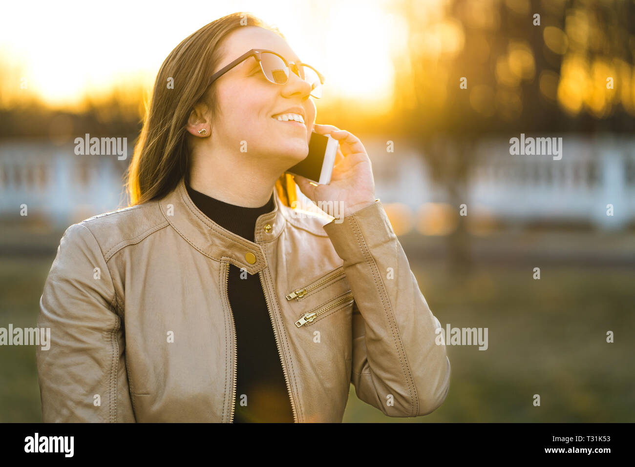 Fröhliche Frau lächelt und lacht, während sie im Freien bei Sonnenuntergang telefoniert. Junge Dame, die ein Gespräch mit einem Freund hat. Person mit Smartphone. Stockfoto