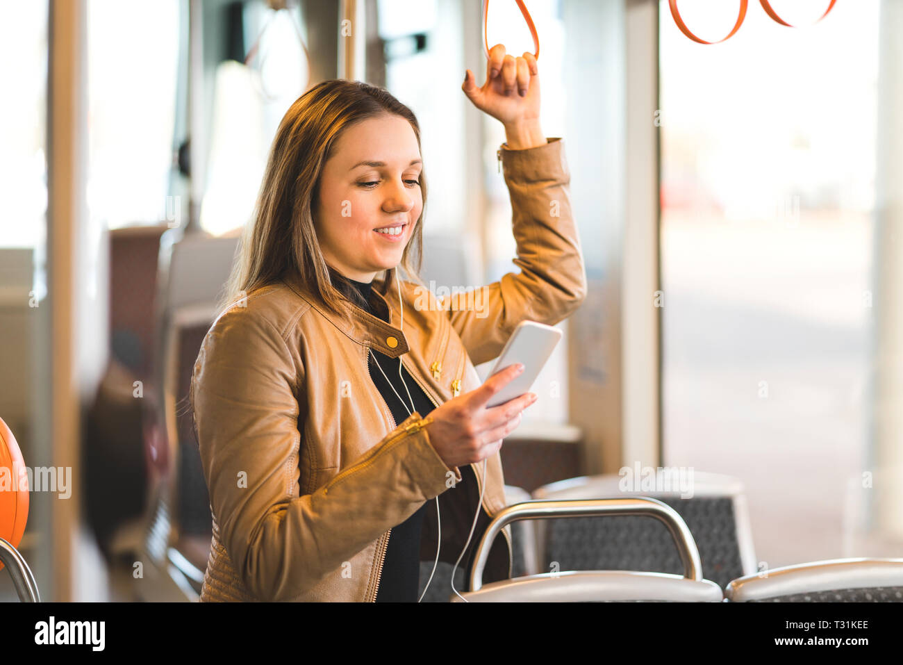Frau, die im Zug, in der Straßenbahn oder im Bus steht, die den Griff hält und das Handy benutzt. Fröhliche weibliche Beifahrerin textet während der Fahrt mit dem Smartphone. Stockfoto