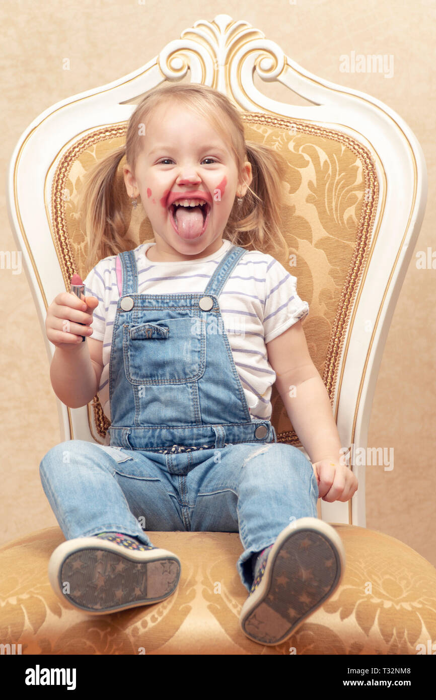 Porträt eines kleines fröhliches Mädchen mit Zunge heraus, mit Lippenstift auf ein retro Stuhl verschmiert Stockfoto