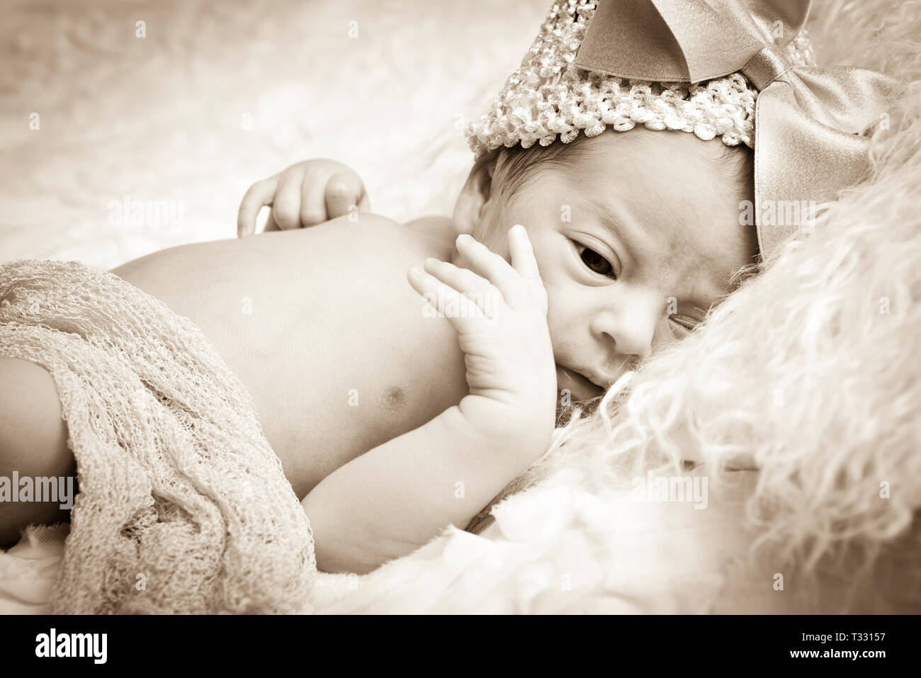 Nahaufnahme einer wachen, schöne neugeborenes Mädchen ruht auf einem Fell Kissen Tragen eines knit Cap mit großen Bogen, und mit einem Netzgewebe in Sepia Ton drapiert Stockfoto