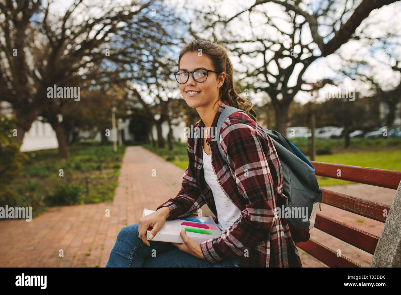 Schöne Schülerin sitzt auf der Bank mit Buch. Frau am Campus der Universität mit Blick auf die Kamera und lächelnd. Stockfoto