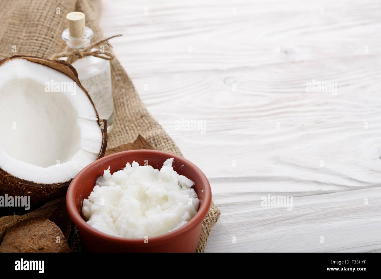 Kokosöl in braun Ton Schale, Glas, Flasche, Kokosnussschalen mit Fleisch auf Hanf Sack auf weißen Küche aus Holz Tisch Stockfoto