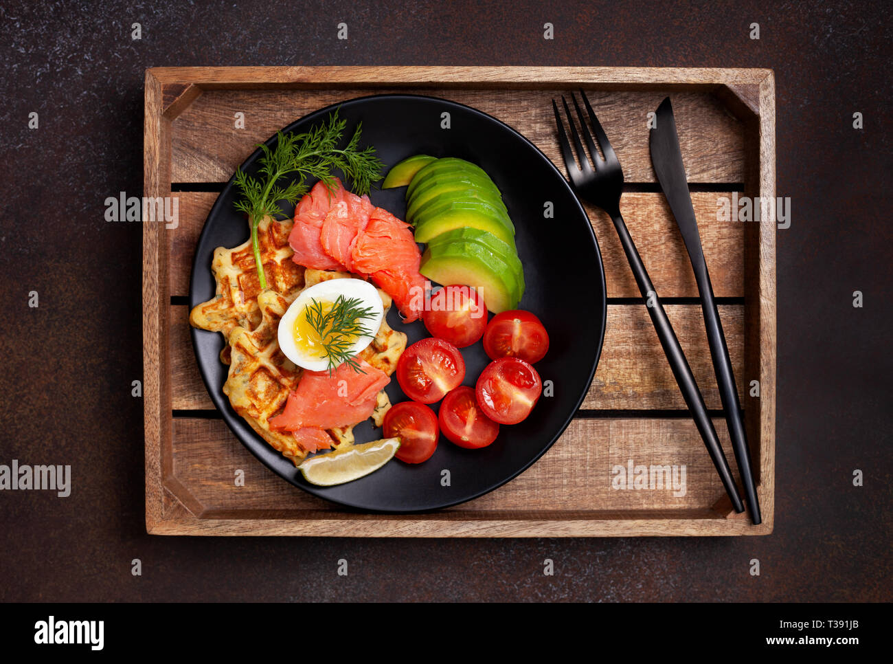 Kartoffel Waffeln mit Lachs, Ei, Avocado und Tomaten auf einer schwarzen Platte auf einem dunklen Hintergrund. Blick von oben. Stockfoto