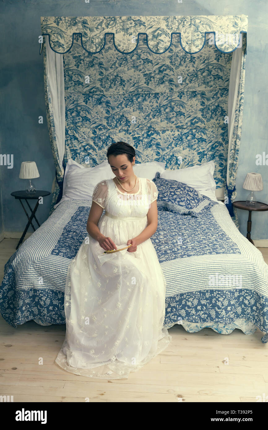 Attraktive junge Frau in authentischen Regency Kleid sitzt auf einem antiken Bett Stockfoto