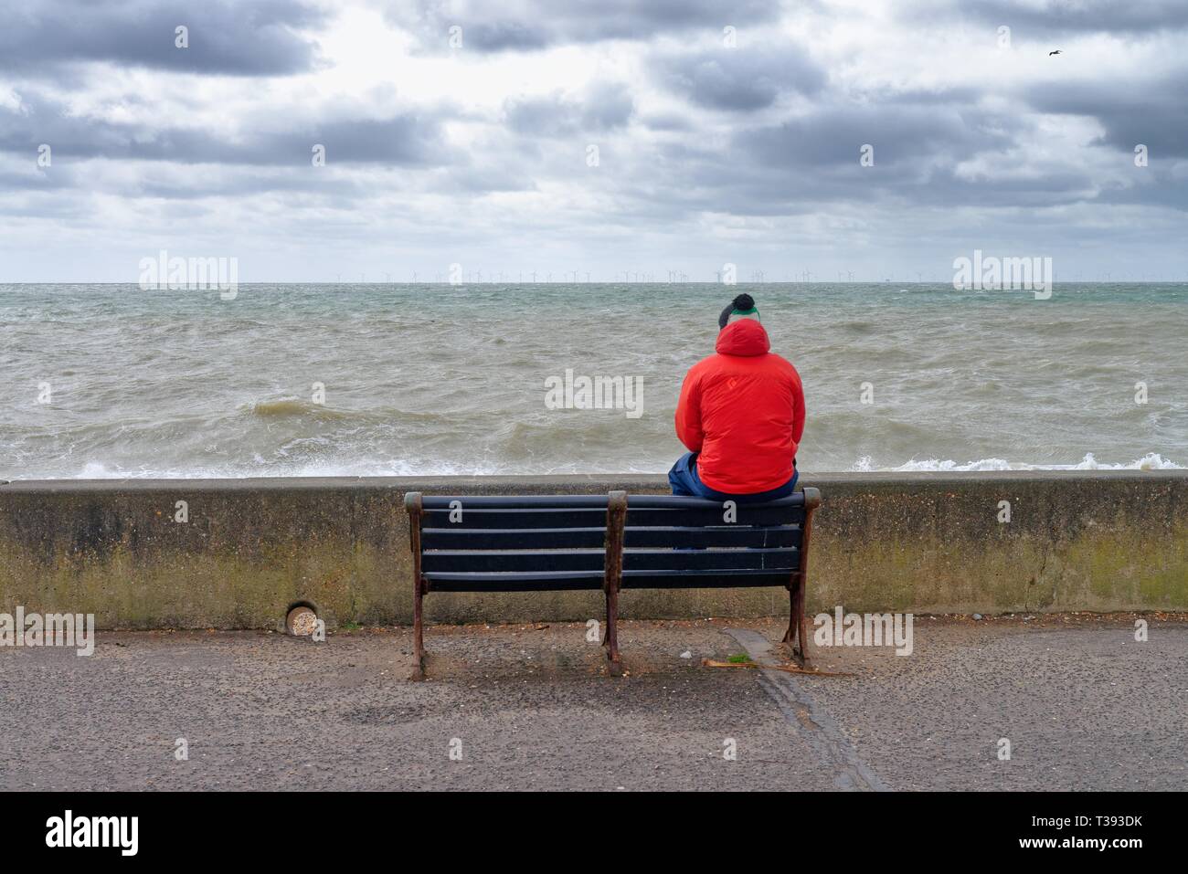 Männliche Figur im roten Mantel sitzt auf der Rückseite der Rücksitzbank aus Blick auf das Meer an der Küste von Hove, East Sussex England Großbritannien Stockfoto