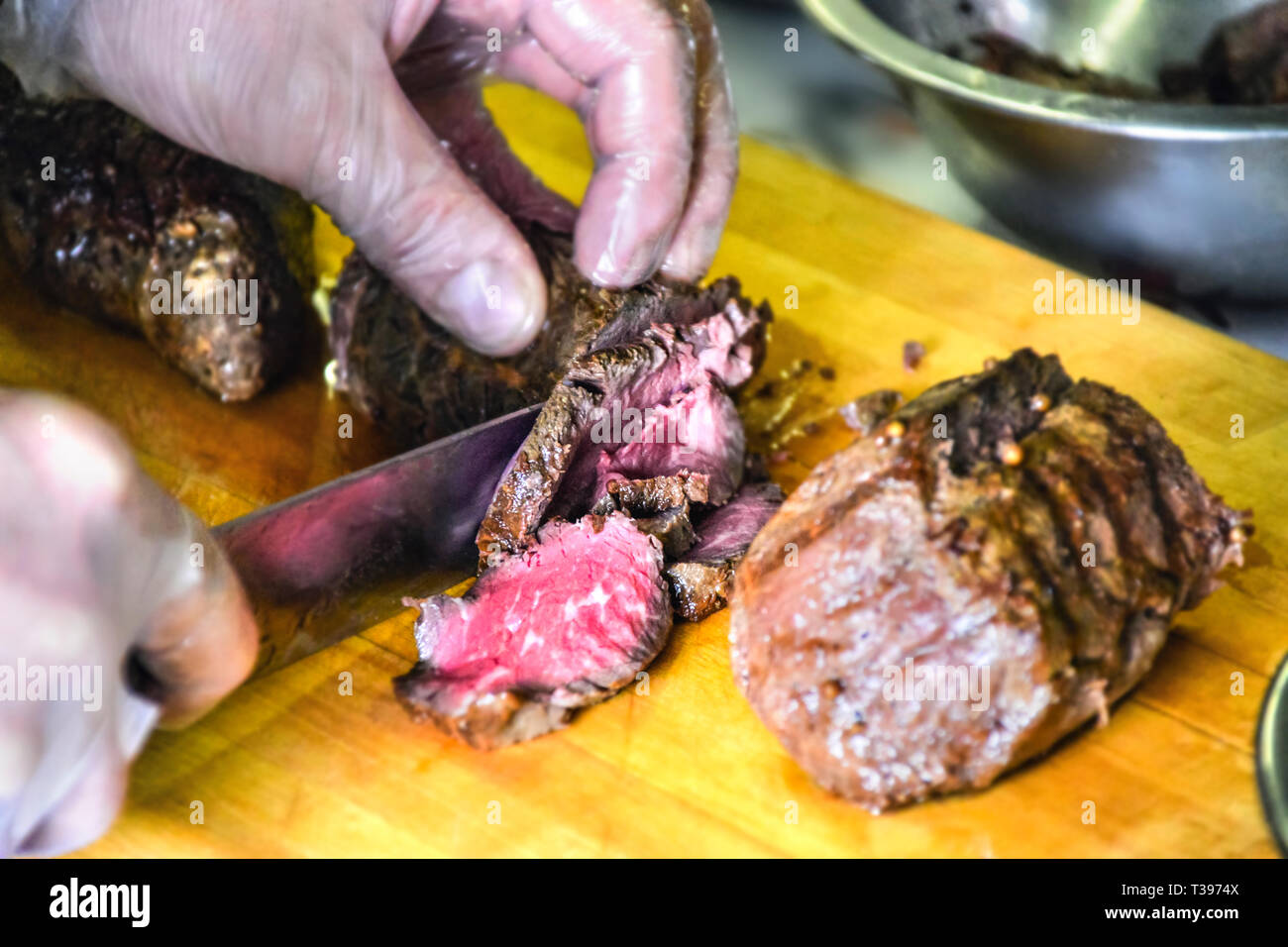 Koch Hände in Einweg Zubereitung Handschuhe aus Kunststoff Carving gekocht Roastbeef Roastbeef mit einem großen Messer auf einer hölzernen Schneidebrett. Catering Servic Stockfoto