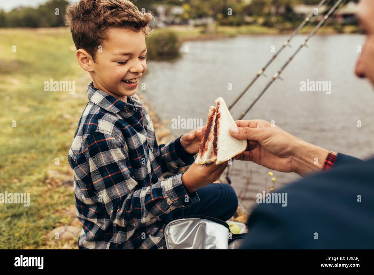 Kind eine Pause vom Angeln Sandwiches zu essen. Mann, Sandwich zu seinem Kind, während neben einem See zum Angeln zu sitzen. Stockfoto