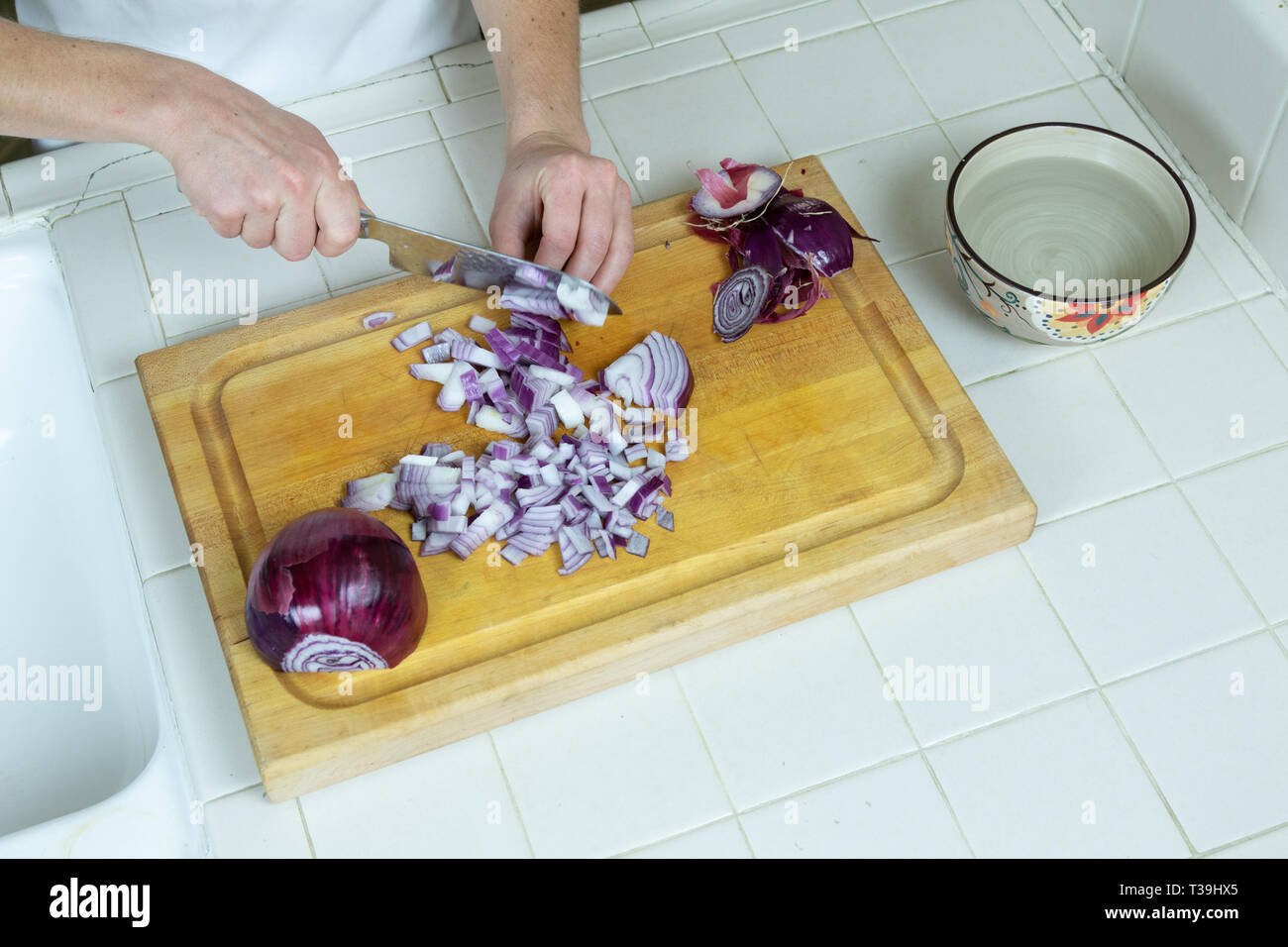 Ansicht von oben der Arbeitsplatte in der Küche, ein Schneidbrett, Schüssel mit Wasser, Ecke von Spülbecken, mit weiblichen Händen würfeln rote Zwiebel. Stockfoto