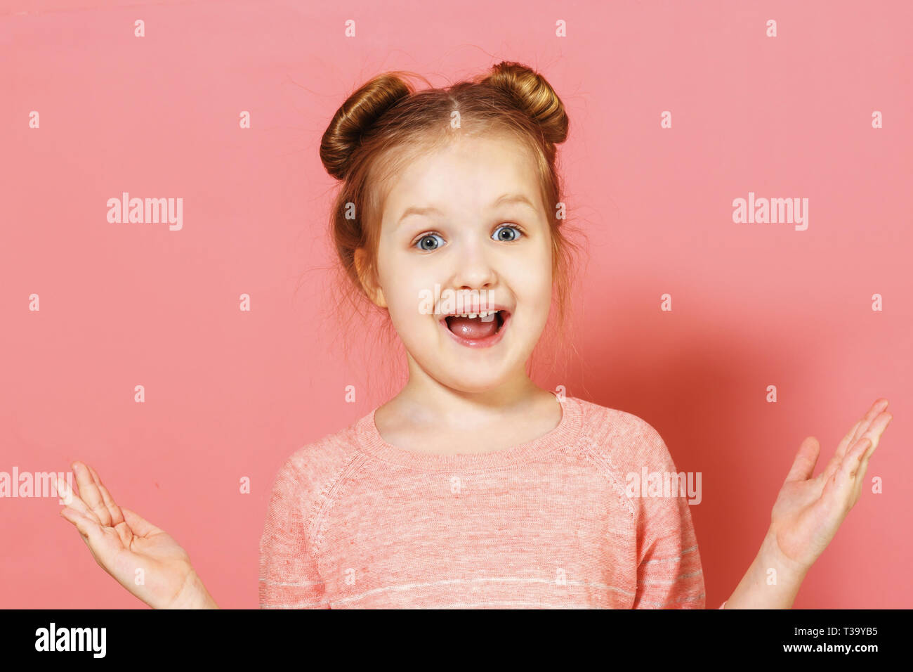 Closeup Portrait von attraktiven charmante fröhliche kleine Mädchen mit Brötchen über rosa Hintergrund. Das Kind in der Überraschung und beide Hände an den Seiten verteilt. Stockfoto