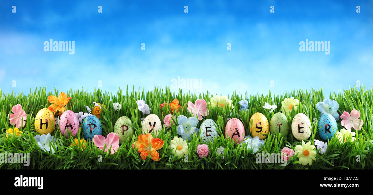 Leuchtend bunte Ostereier sagen Frohe Ostern auf grünem Gras mit Blumen gegen den blauen Himmel Stockfoto