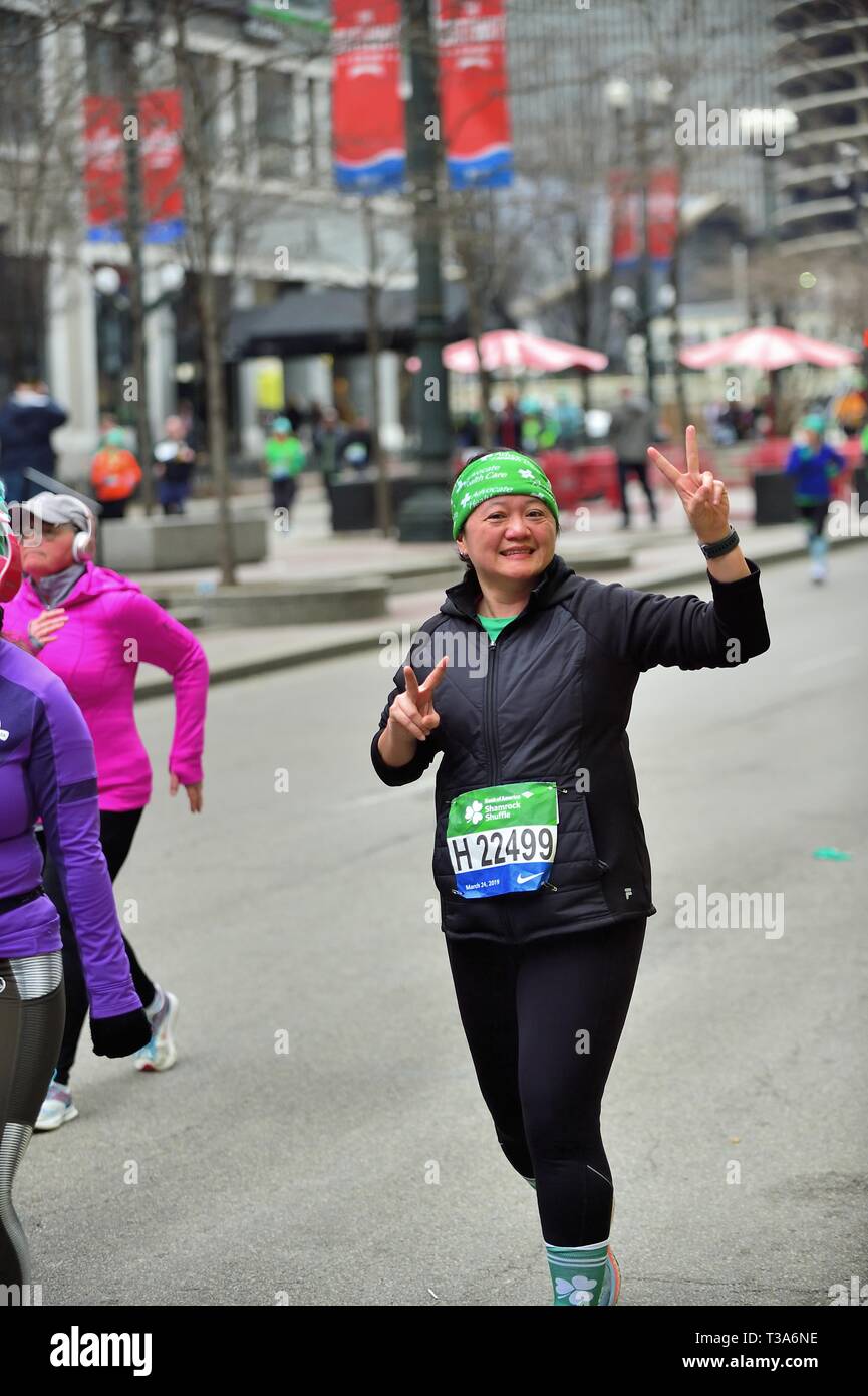 Chicago, Illinois, USA. Eine Frau, die Fortschritte bei der State Street 2019 Shamrock Shuffle Rennen blinkt ein Sieg unterzeichnen ein Verfechter in der Masse. Stockfoto