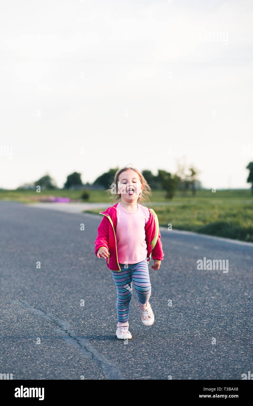 Kleine niedliche Mädchen Spaß haben laufen auf der Straße, Ihre Zunge heraus zu haften, Spielen im Freien Stockfoto