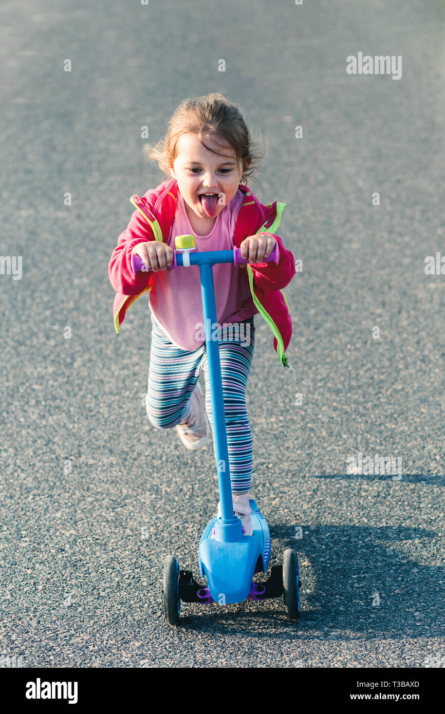 Kleine niedliche Mädchen Spaß reiten auf Motorroller, ihr die Zunge heraus, während die Beschleunigung von Scooter, Spielen im Freien Stockfoto